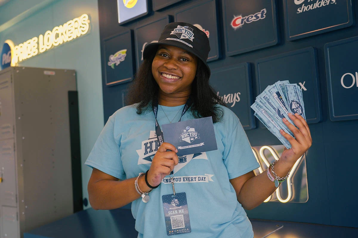 A young, black female college student holds up a blue card and P&G prize money. She is wearing a light blue t-shirt.