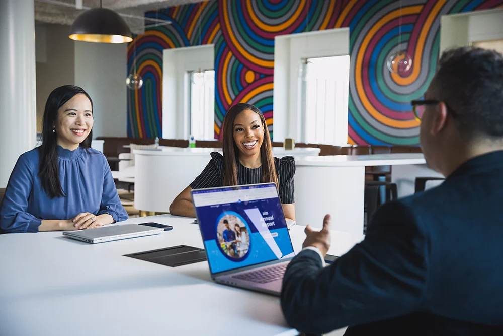 Two women seated at a table. They smile at a man across the table. He has a laptop open in front of him.