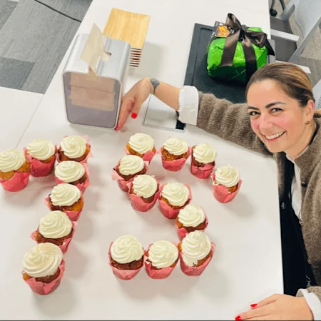 A woman in a brown sweater sits at white office table. In front of her are eighteen cupcakes arranged in the number 15.