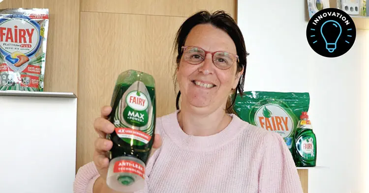 Woman with dark hair and glasses holds up a bottle of green Fairy brand dish soap. A similar bottle is behind her, as well as a larger green bag of Fairy brand dishwasher soap tablets. A large silver bag of Fairy brand soap tablets is placed behind her.