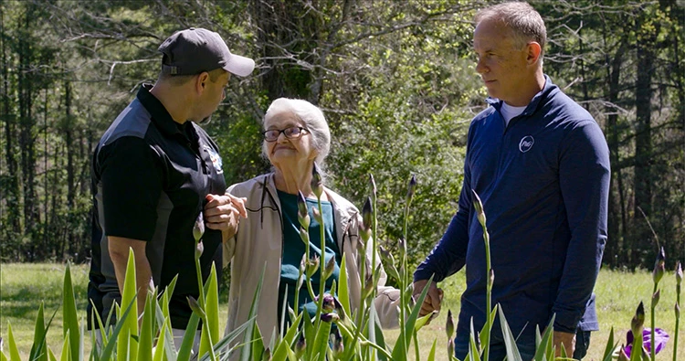Two men and an elderly woman hold hands while they stand outside in front of a patch of long stemmed purple flowers. Numerous trees are lined up across the background.