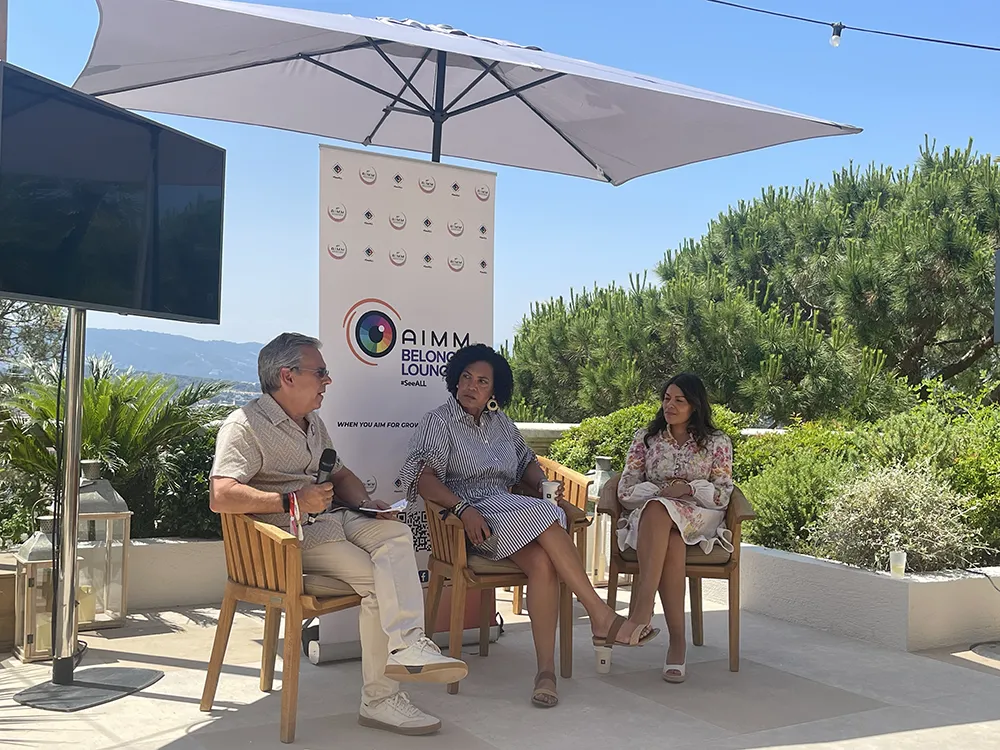 A man and two women sit on a shaded stage, holding microphones and exchanging in dialogue.