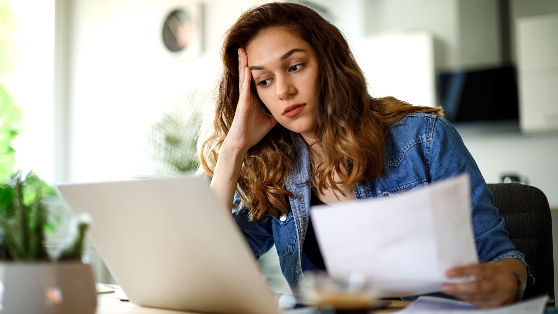 Exasperated looking young woman sitting at desk looking at laptop holding papers in one hand and her head in the other