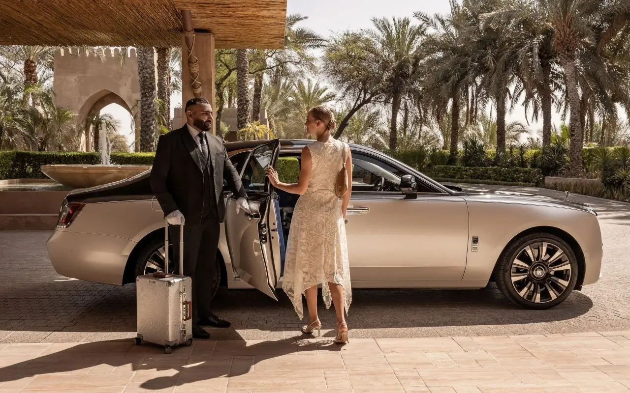 An elegant woman is helped by a chauffeur into a Blacklane Rolls Royce Phantom limousine outside a fancy hotel in Dubai.