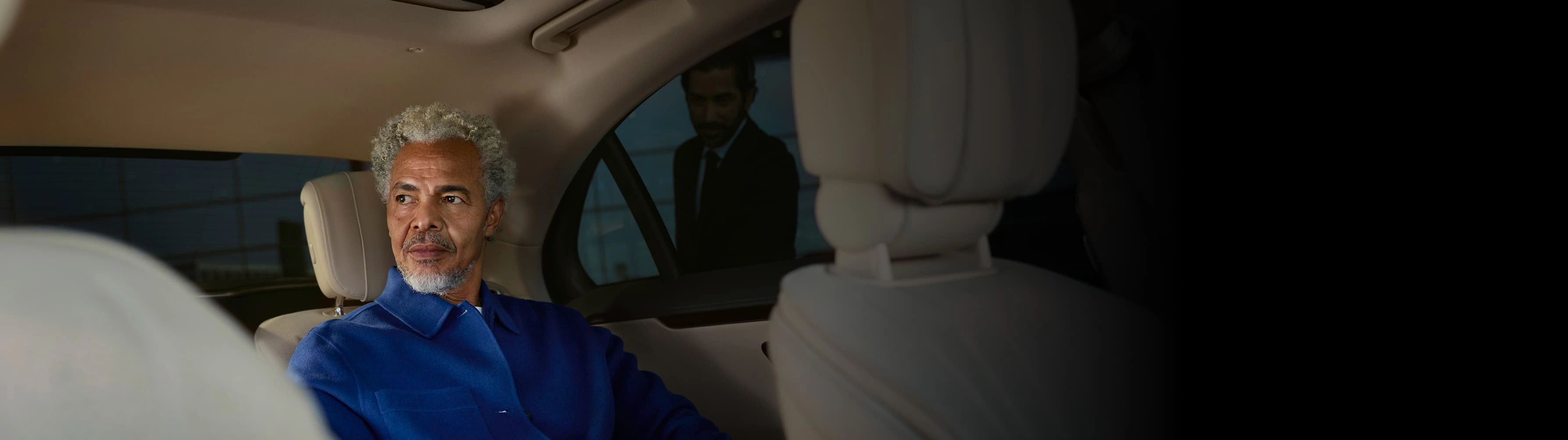 A man sits in the back seat of a Blacklane limo as a chauffeur waits to open the door outside.