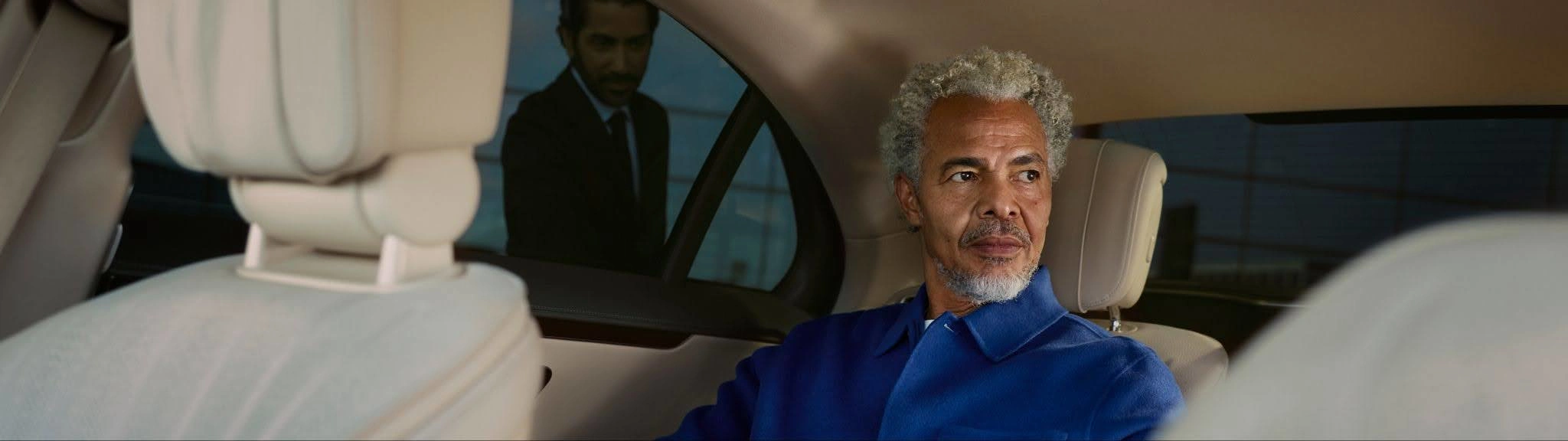 A man sits in the back seat of a Blacklane limo as a chauffeur waits to open the door outside.