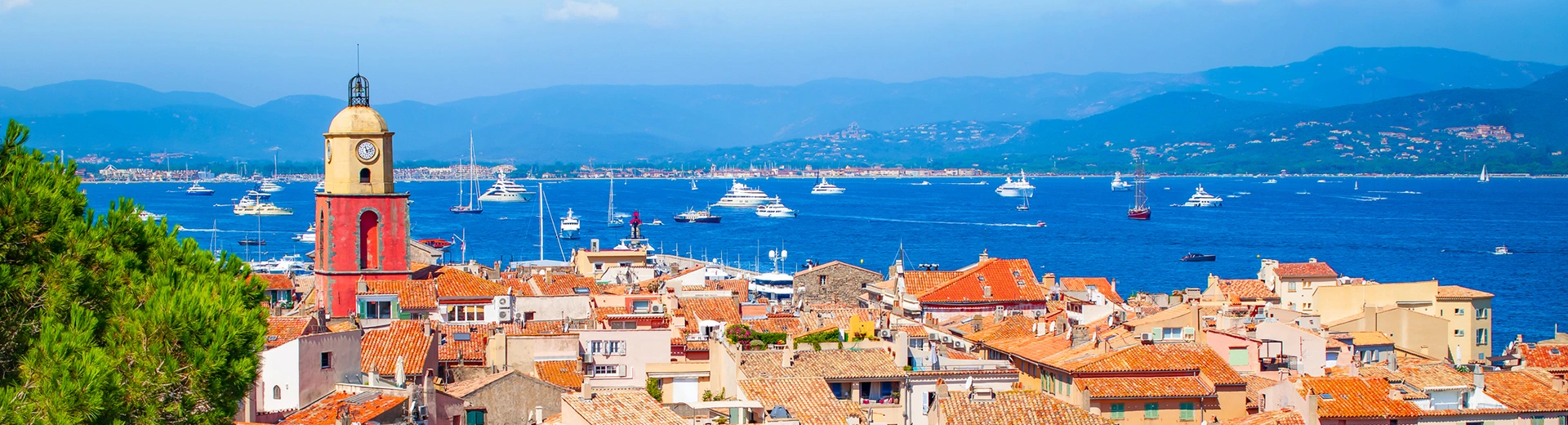 Une vue sur le port de Saint Tropez avec un clocher d'église et des toits au premier plan et de l'eau bleue parsemée de bateaux en arrière-plan.