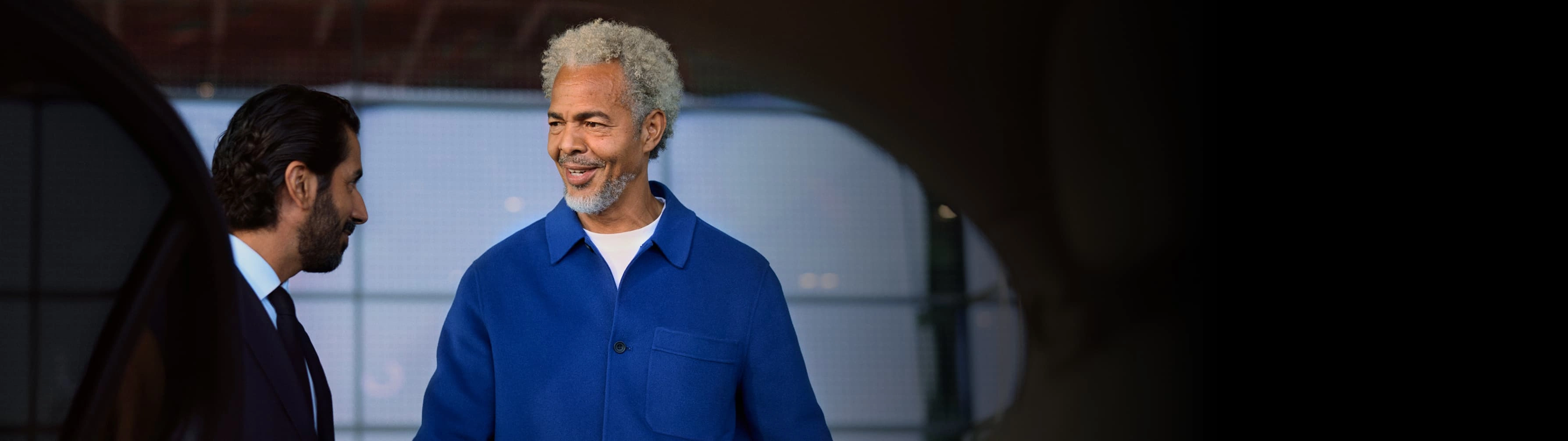 A traveller with grey hair greets his chauffeur while getting into a car outside an airport.