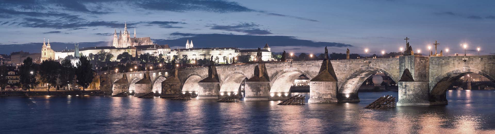 The famous Charles Bridge in Prague is deftly illuminated under a sky of dusk or dawn, with historic spires in the background.
