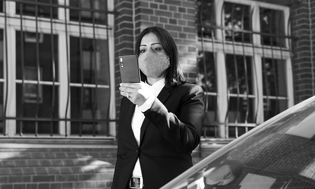 A woman in a suit and face mask stands outside, looking at her phone.