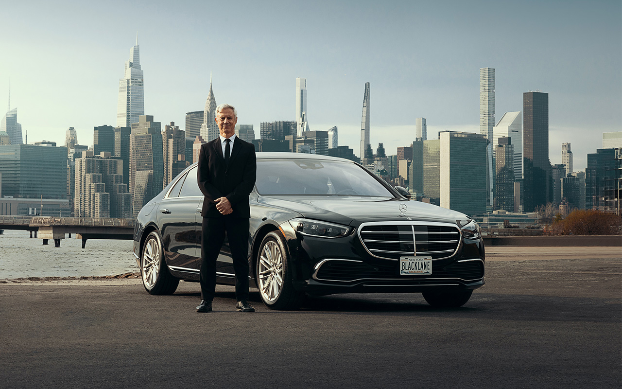 A chauffeur in a suit stand in front of his Mercedes-Benz with the city skyline behind.