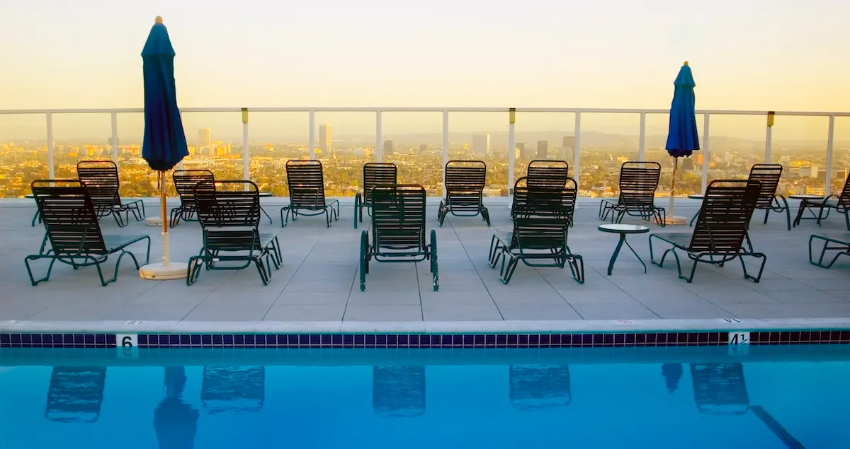 A rooftop pool with sun loungers looking out at a smoggy Los Angeles lit by a sunset.