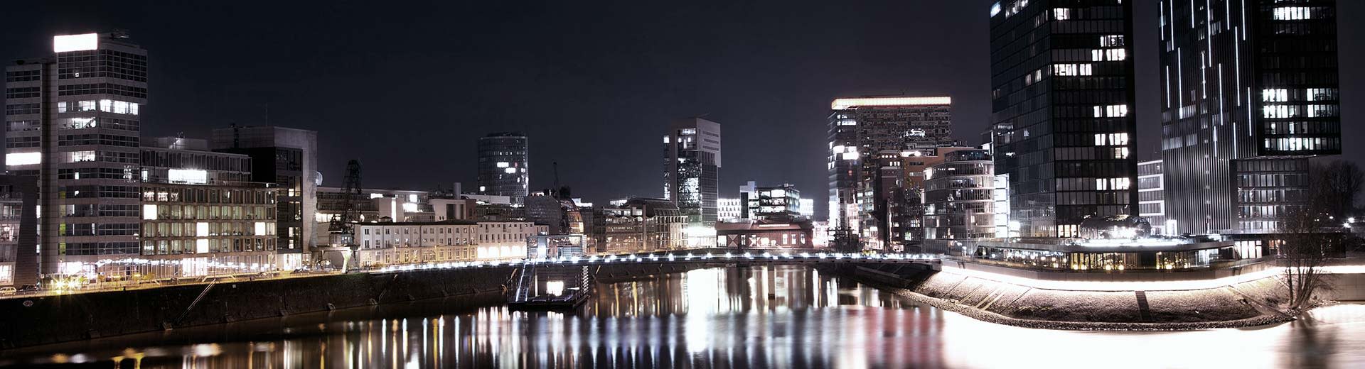 A bridge over a body of water in Neuss at night, with tall buildings lighting up the immediate area.