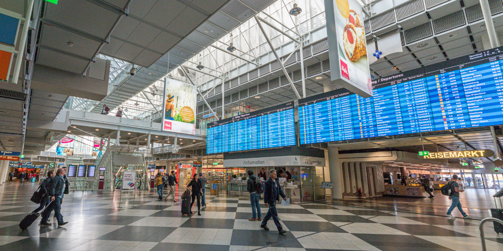 Travelers walking through a modern terminal at Munich Airport, with digital flight information boards and bright signage above.