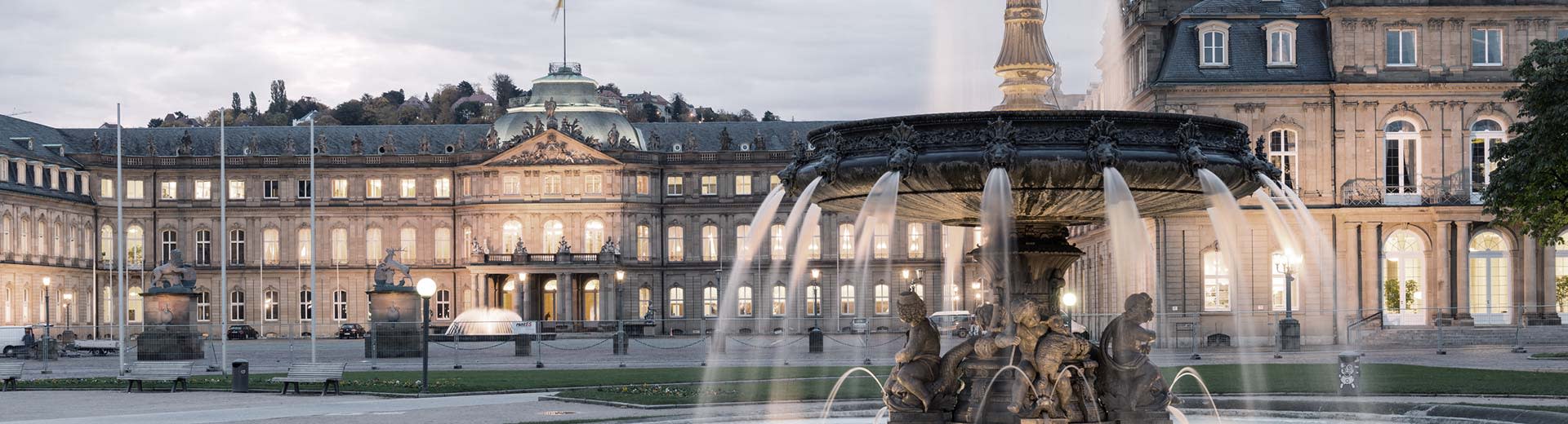 A beautiful fountain dominates the foreground, while behind stands a historic building in the half-light of a Stuttgart evening.