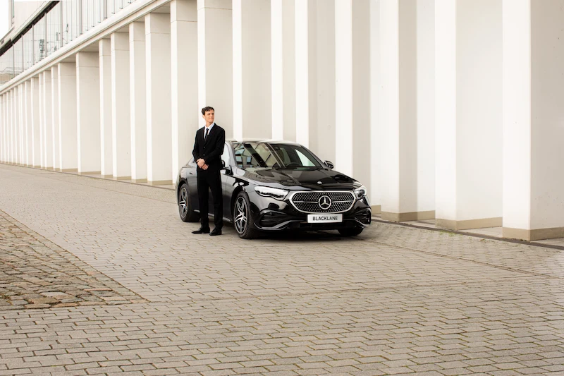 A suited Blacklane chauffeur stands beside a black Mercedes E-Class limo next to a building in Berlin's Mitte.