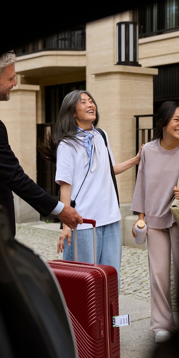 A Blacklane chauffeur takes a passenger's suitcase as she and her daughter laugh.
