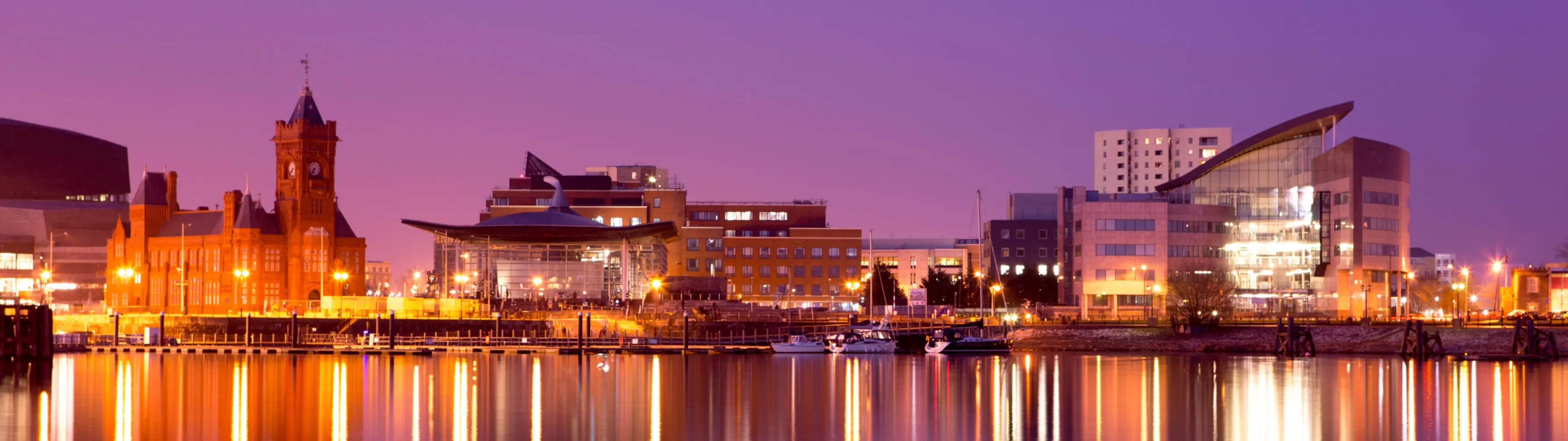 The skyline of Cardiff at night, reflected in the dark water that lies before it.