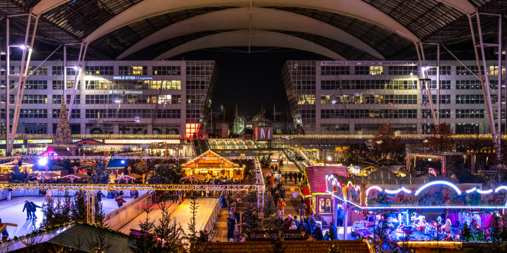 Festive Christmas market and ice rink set up at Munich Airport, illuminated at night under the airport’s large glass roof.