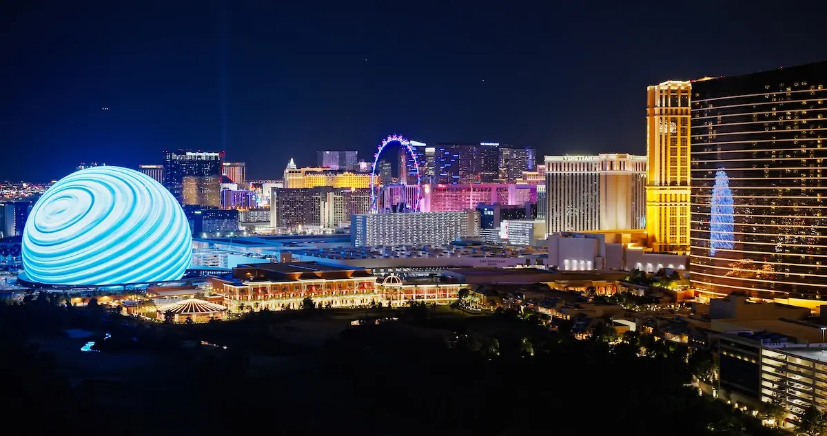 Shot of the Las Vegas skyline, including the Bellagio and the Sphere all lit up in the night sky, with a Ferris Wheel in the background.