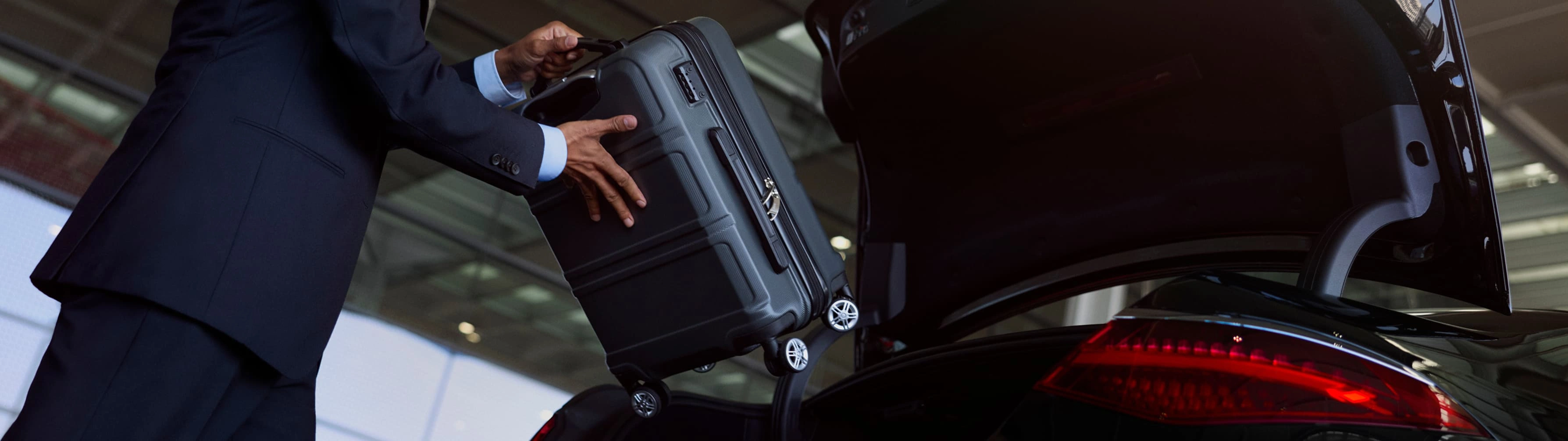 A suited Blacklane chauffeur places a luggage case in the back seat of a black Mercedes luxury vehicle.