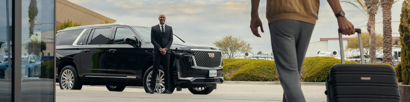 Ein Blacklane Chauffeur steht neben seiner Cadillac Escalade und lächelt einen Gast an, der sich mit einem rollenden Koffer nähert.