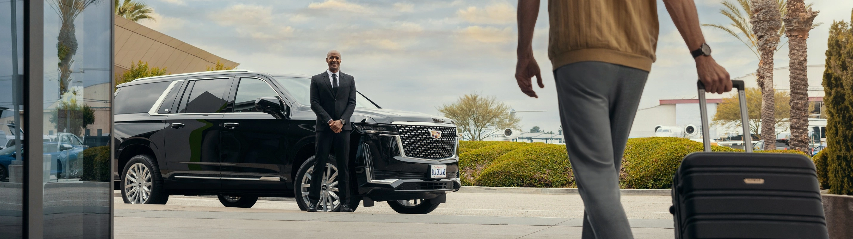 A Blacklane chauffeur stands next to his Cadillac Escalade, smiling at a guest who approaches with a rolling suitcase.