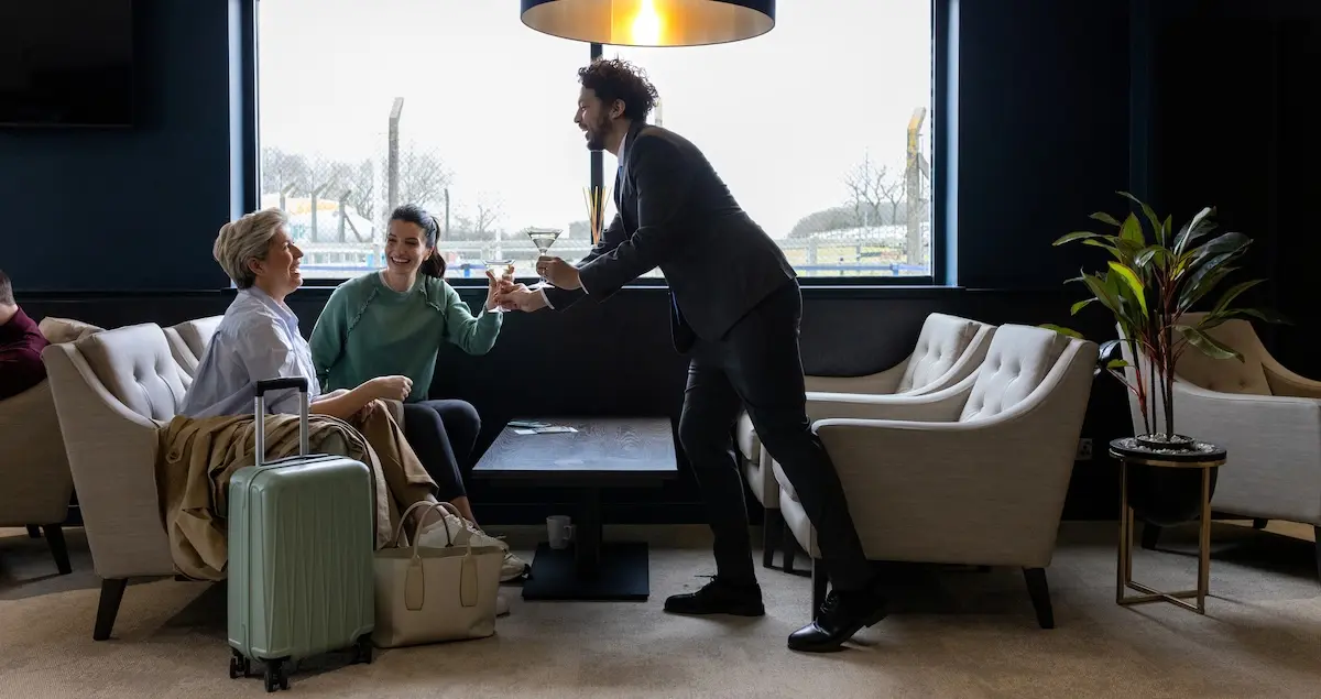 A full shot of a male waiter handing cocktails to two women sitting with their baggage in an airport VIP lounge.