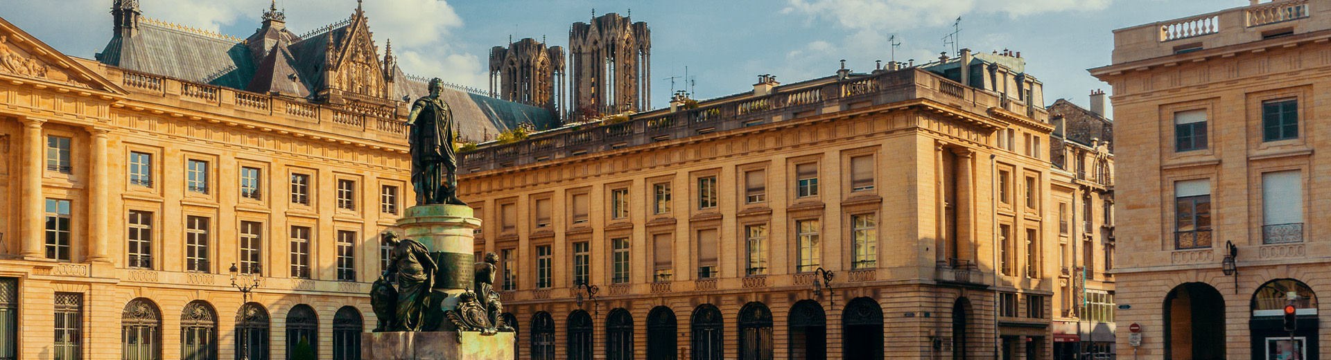 Una imagen de Place Royale, una plaza en Reims. En primer plano se encuentra una estatua de bronce de Louis XV. Las torres gemelas de Cathédale Notre-Dame son visibles por encima de los edificios en el fondo.