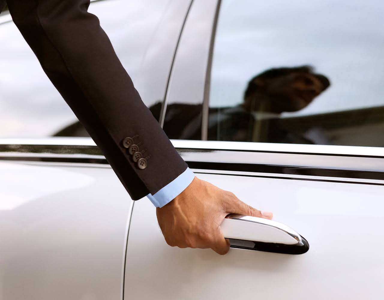 Close-up of a Blacklane chauffeur's suited arm holding the door handle of a two-tone luxury car.