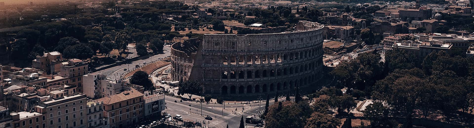 In the half-light of dusk or dawn, the famous Colosseum of Rome dominates the image, surrounded by buildings.