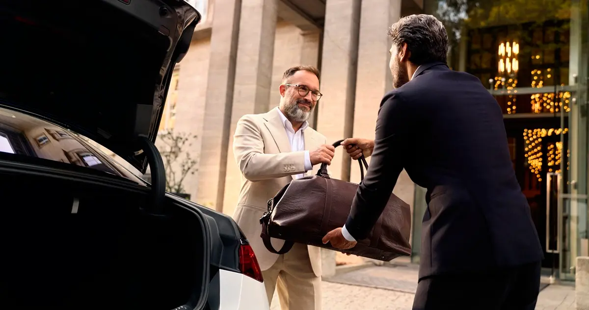 A smartly-dressed man outside a private members' club takes his bag from a chauffeur with an open trunk in the foreground.