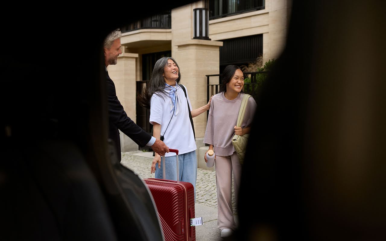 A woman and daughter stand laughing with luggage beside a suited Blacklane chauffeur who is taking their bags to the car.