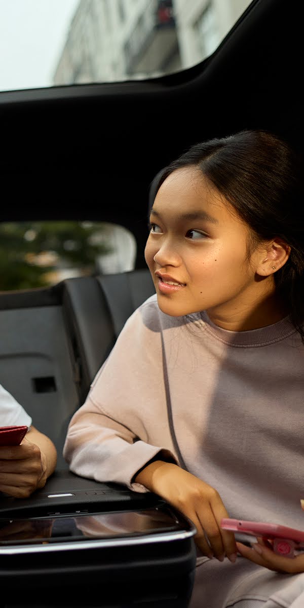 A young girl gazes out the backseat window of a luxury car in wonder which holding her smartphone.