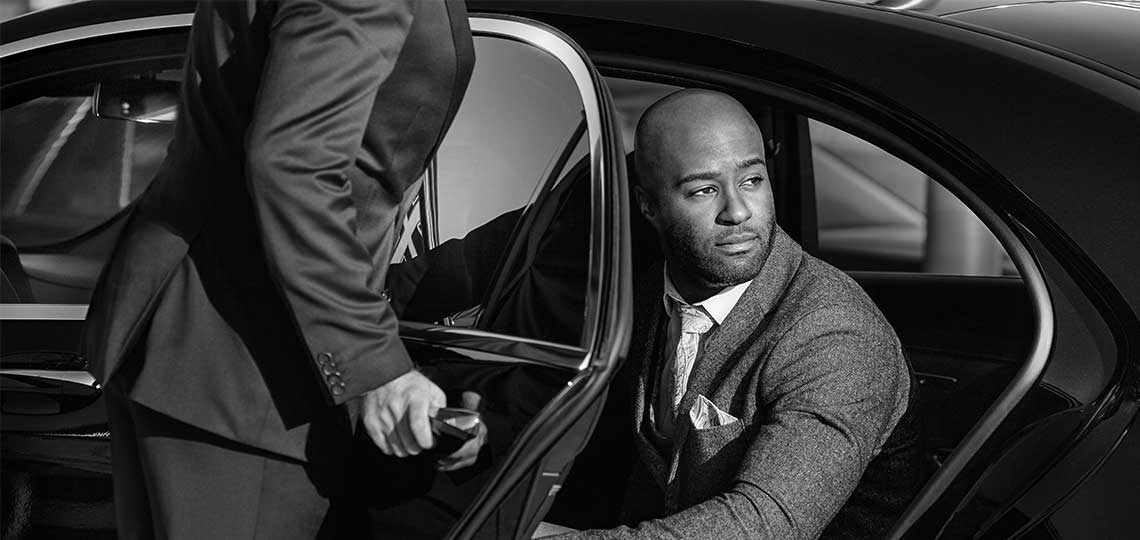 A Blacklane chauffeur in a black suit opens the door to let a business traveler in a grey suit out of the vehicle.