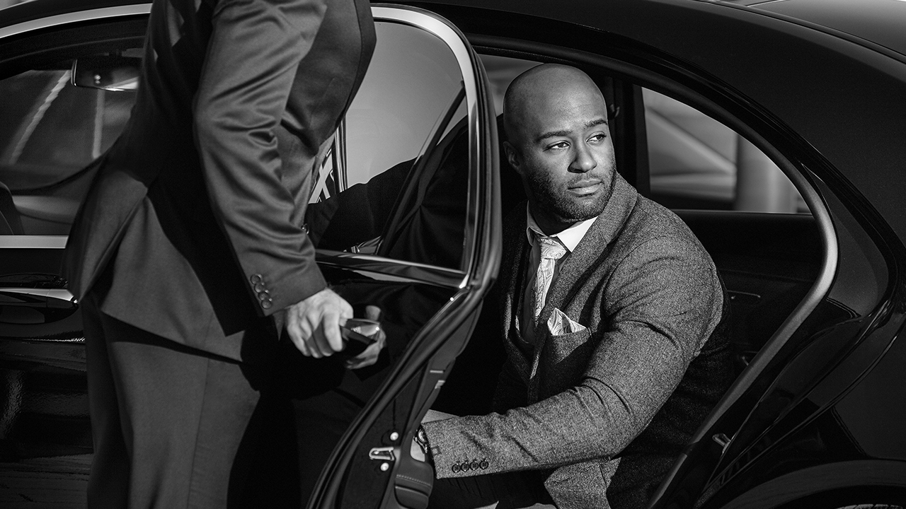 A Blacklane chauffeur in a black suit opens the door to let a business traveler in a grey suit out of the vehicle.