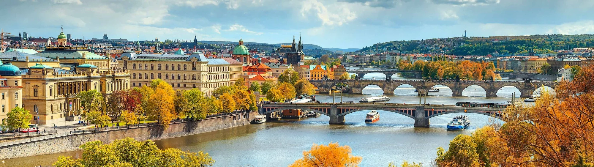 The Czech city of Prague shot from the riverbank looking over the city's bridges with autumnal trees everywhere.