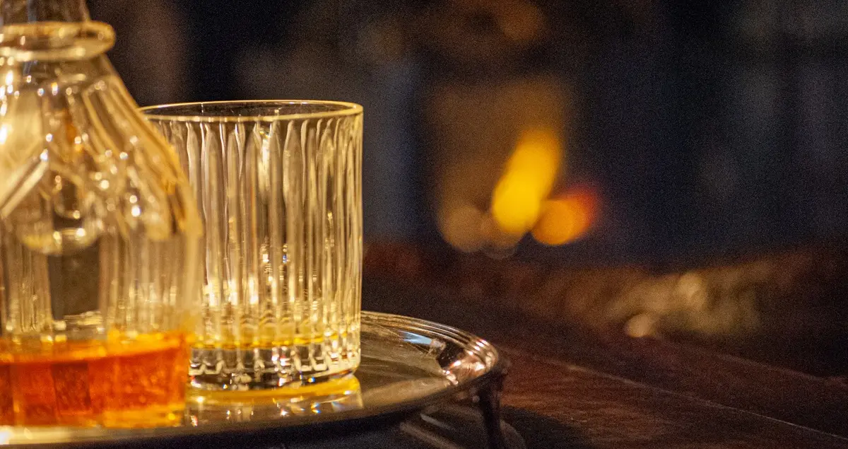 Close-up of a whiskey tumbler with a decanter next to it on a silver platter with a fire in a fireplace in the background.