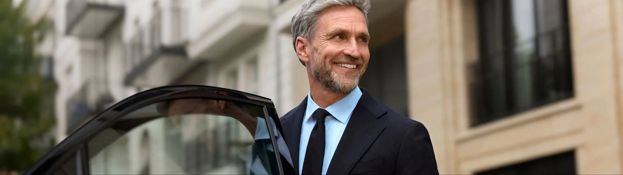A Blacklane chauffeur in a dark suit stands smiling and holding open the door of his Blacklane limousine.