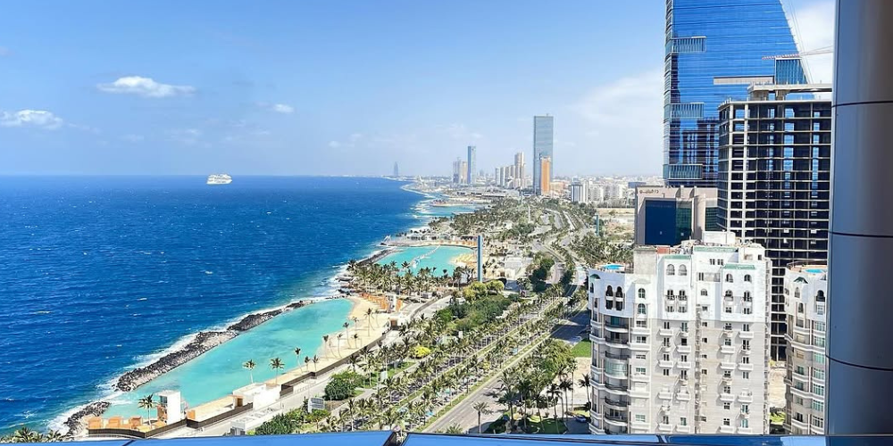 A scenic view of Jeddah’s coastline with high-rise buildings, palm-lined roads, and the Red Sea on a sunny day.