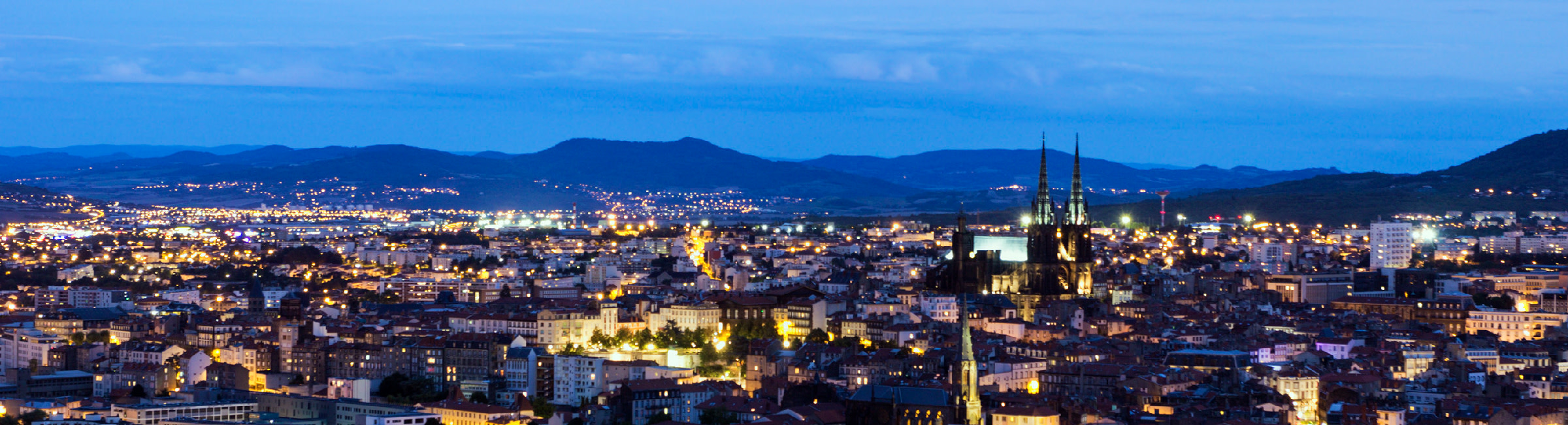 An ariel view, at night, of the cosy French town of Clermont Ferrand. Old buildings and yellow light lie before distant hills.