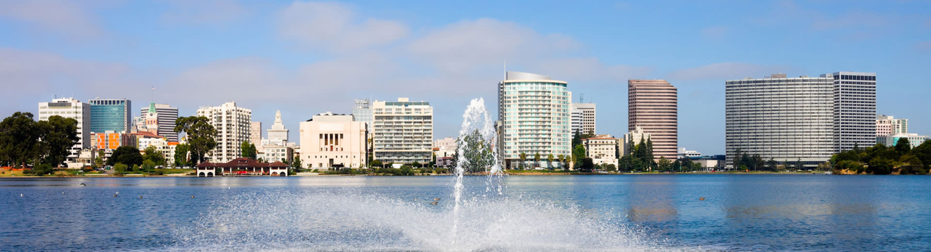 The skyscrapers of Oakland sit behind a body of water, with a fountain in the forgeround.