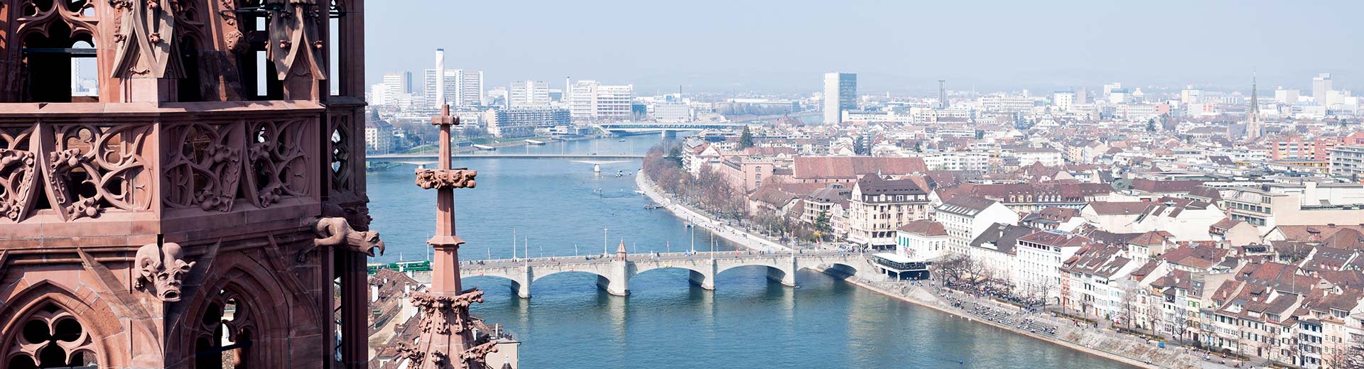 Birds eye view of the Rheine river in Basel on a sunny day, with a tower partially in view on the left and three bridges that cross the river.