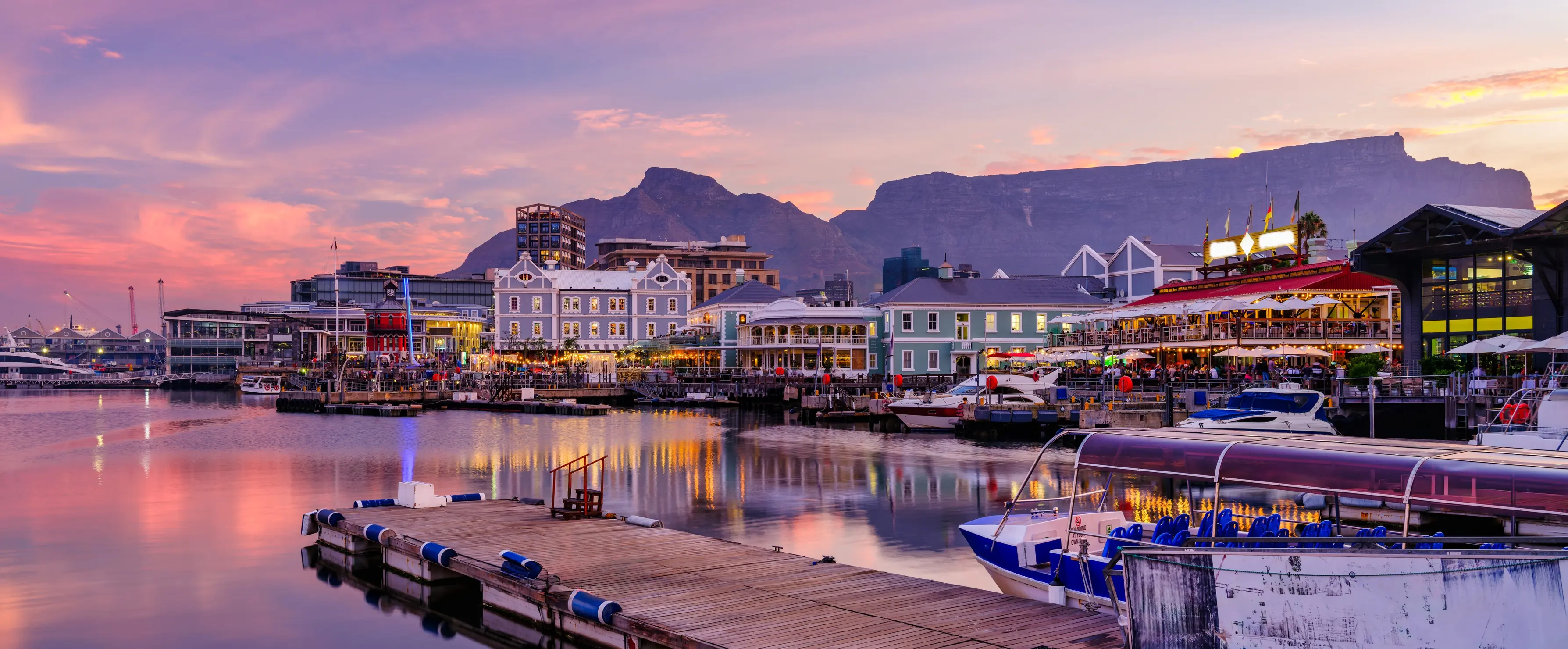 Shot of the V&A Waterfront in Cape Town lit up in purple and violet by a sunset, with Table Mountain in the background.