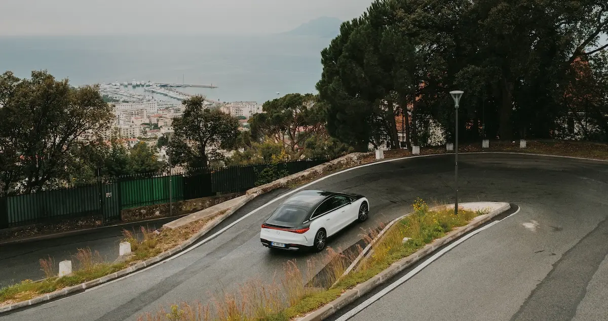 A Blacklane two-tone Mercedes limo travels up a hairpin bend on the Cote d'Azur with a city and harbour in the background.