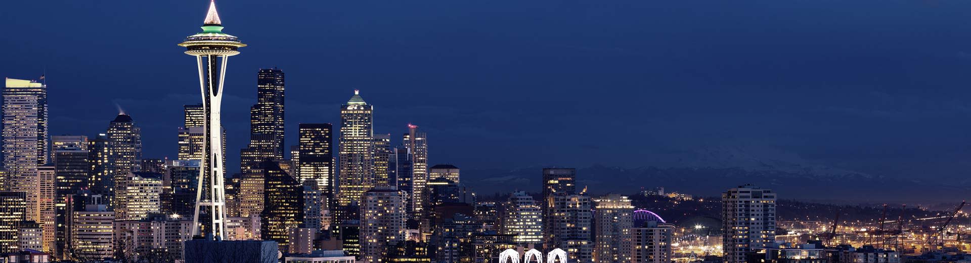 The famous Space Needle of Seattle looms into the nightsky, surrounded by omposing skyscrapers and apartment buildings.
