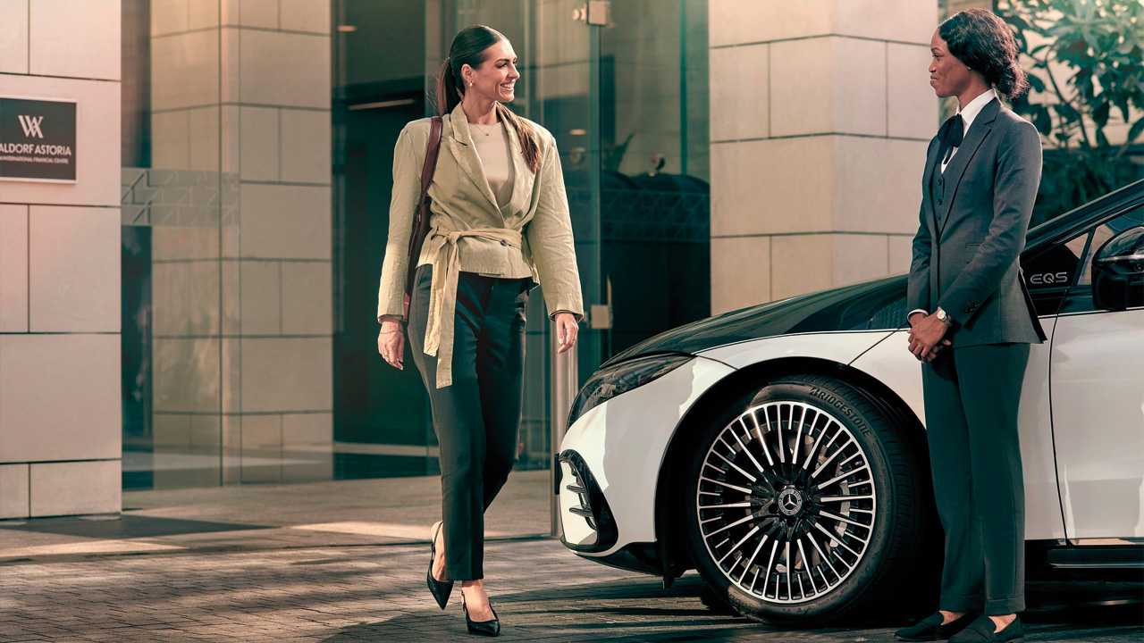 A guest walks toward her chauffeur who stands next to a Mercedes-Benz EQS.