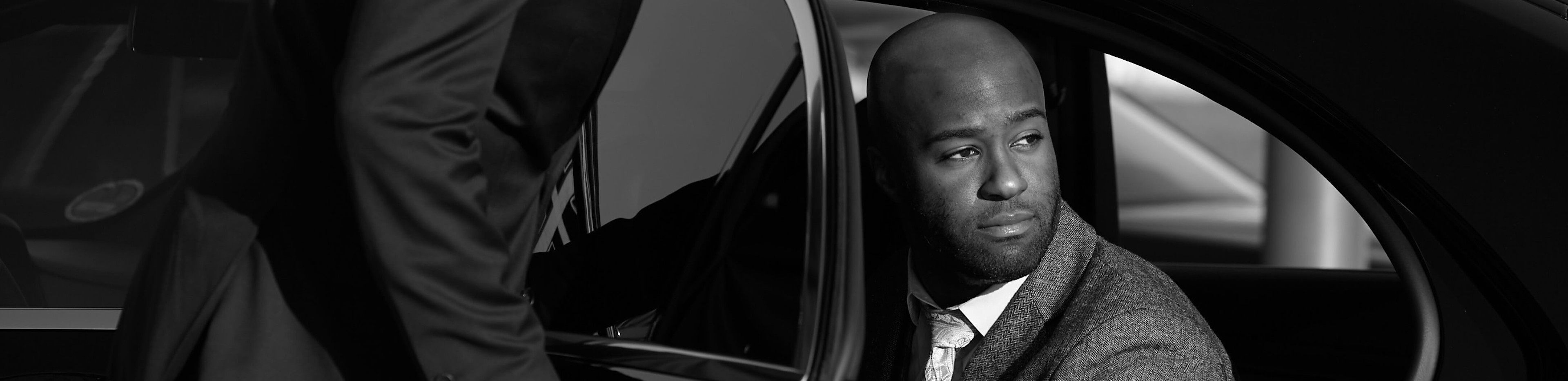 A Blacklane chauffeur in a black suit opens the door to let a man in a grey suit out of the vehicle.