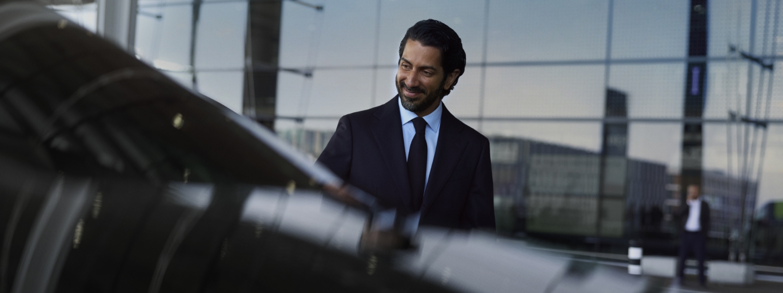 A chauffeur in a suit stands smiling next to a black vehicle.