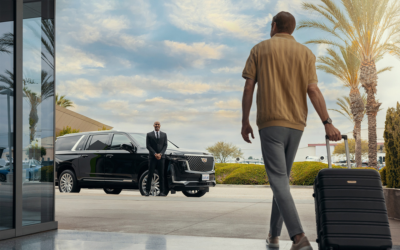 Guest's back is turned to the camera and is walking with rolling luggage towards a Blacklane vehicle while the chauffeur stands ready to greet.